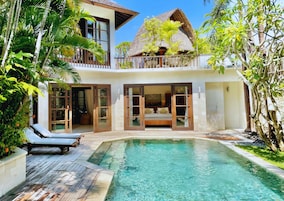 Uma villa de dois andares com piscina, varanda com telhado de palha e vista para vegetação exuberante.