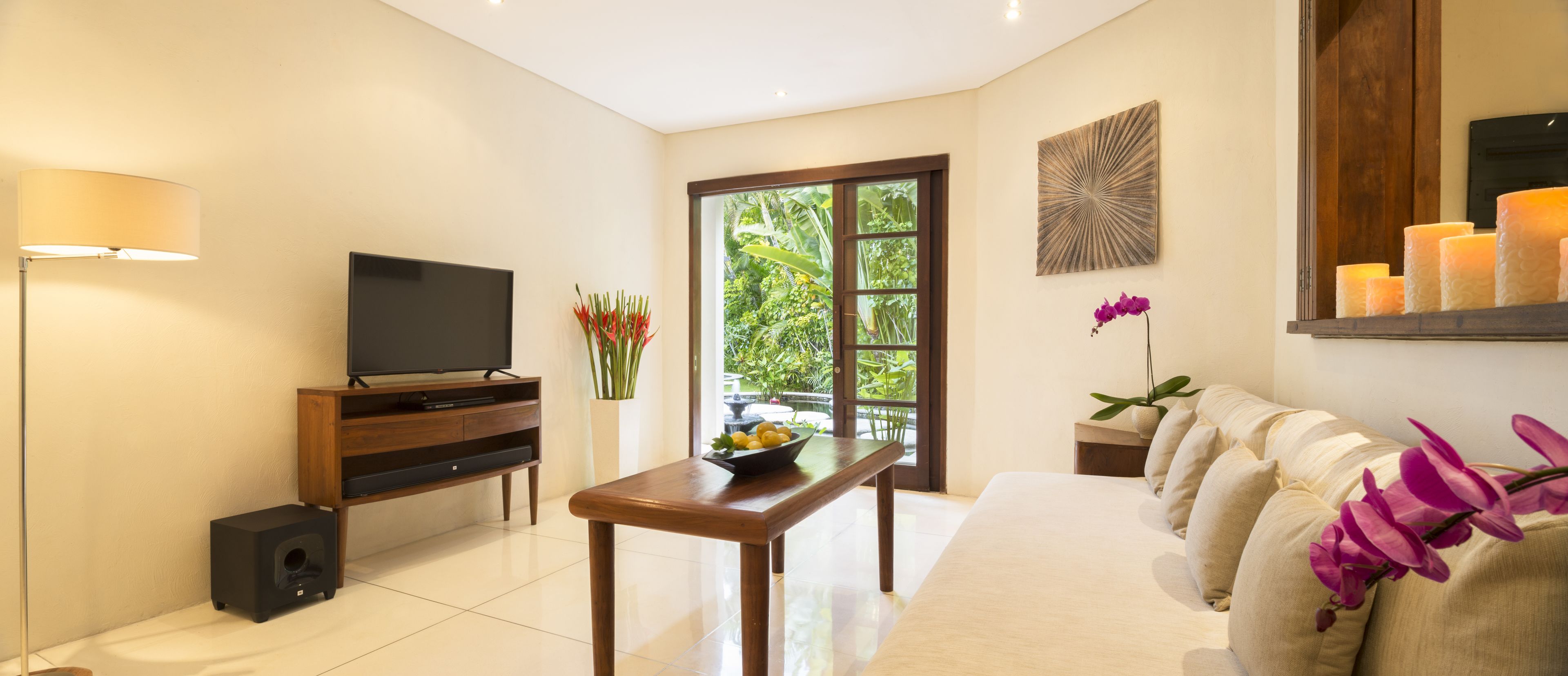 Una sala de estar moderna con un televisor de pantalla plana, un mueble bajo para equipo audiovisual, una mesa de comedor con un cuenco de fruta, un sofá con cojines y un ventanal con vistas a zonas verdes.