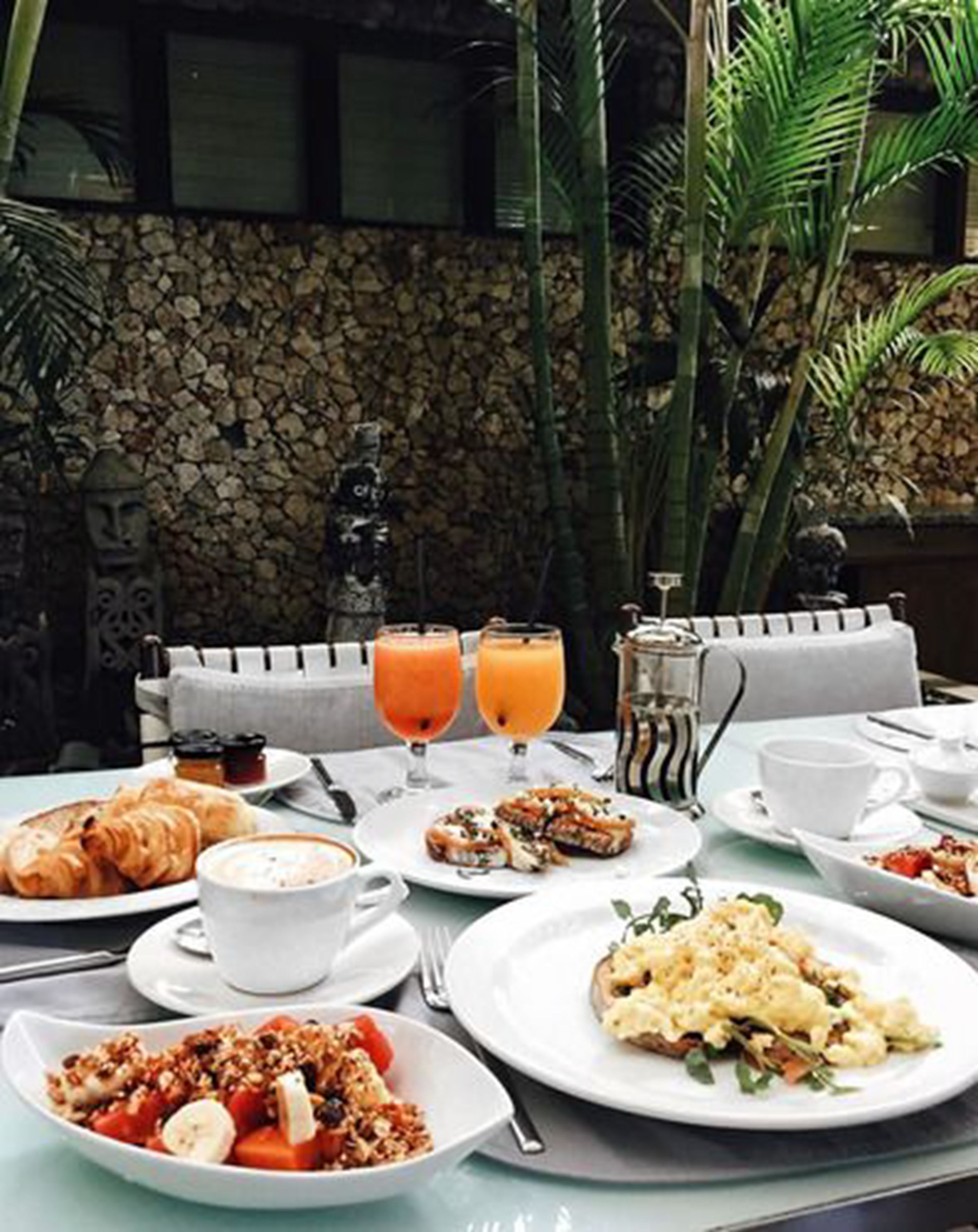 A breakfast setting with croissants, fruit, and coffee on a table with a stone wall backdrop.