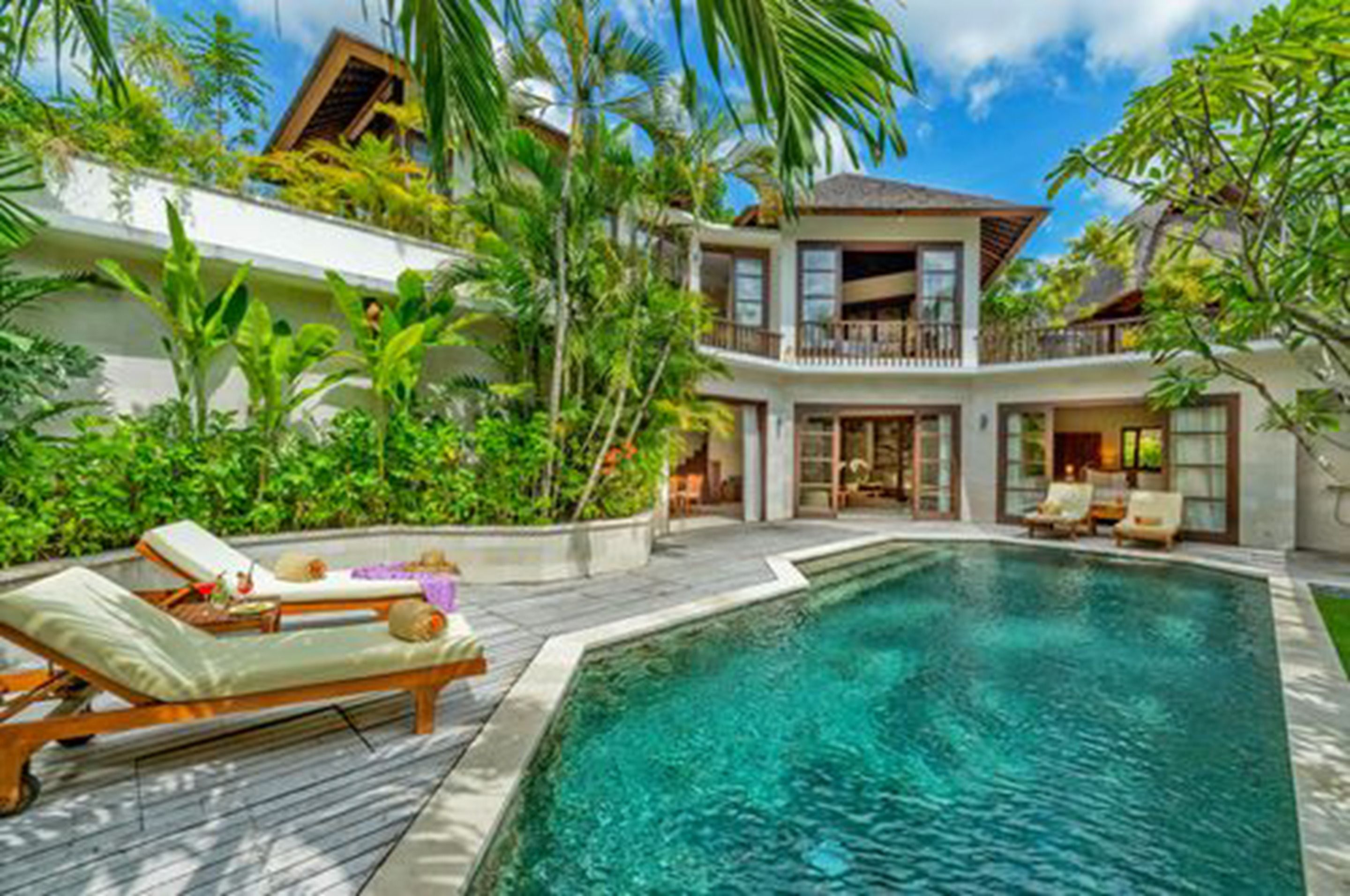 A two-story resort with a swimming pool, lush greenery, and a balcony with lounge chairs.