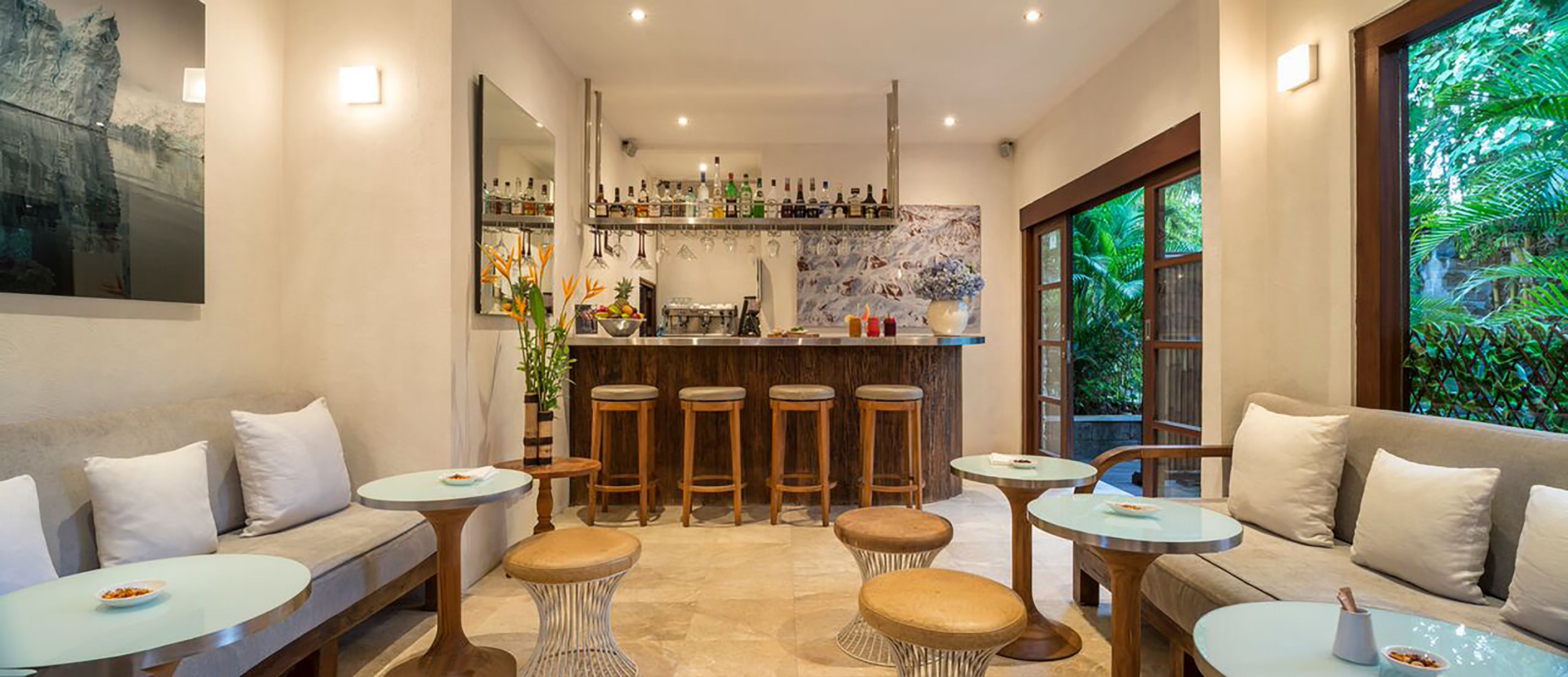 Un área de bar moderna con taburetes, una barra y una variedad de botellas. Hay zonas de estar con mesas redondas y bancos acolchados.