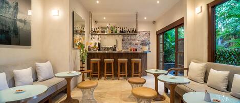 Un área de bar moderna con taburetes, una barra y una variedad de botellas. Hay zonas de estar con mesas redondas y bancos acolchados.