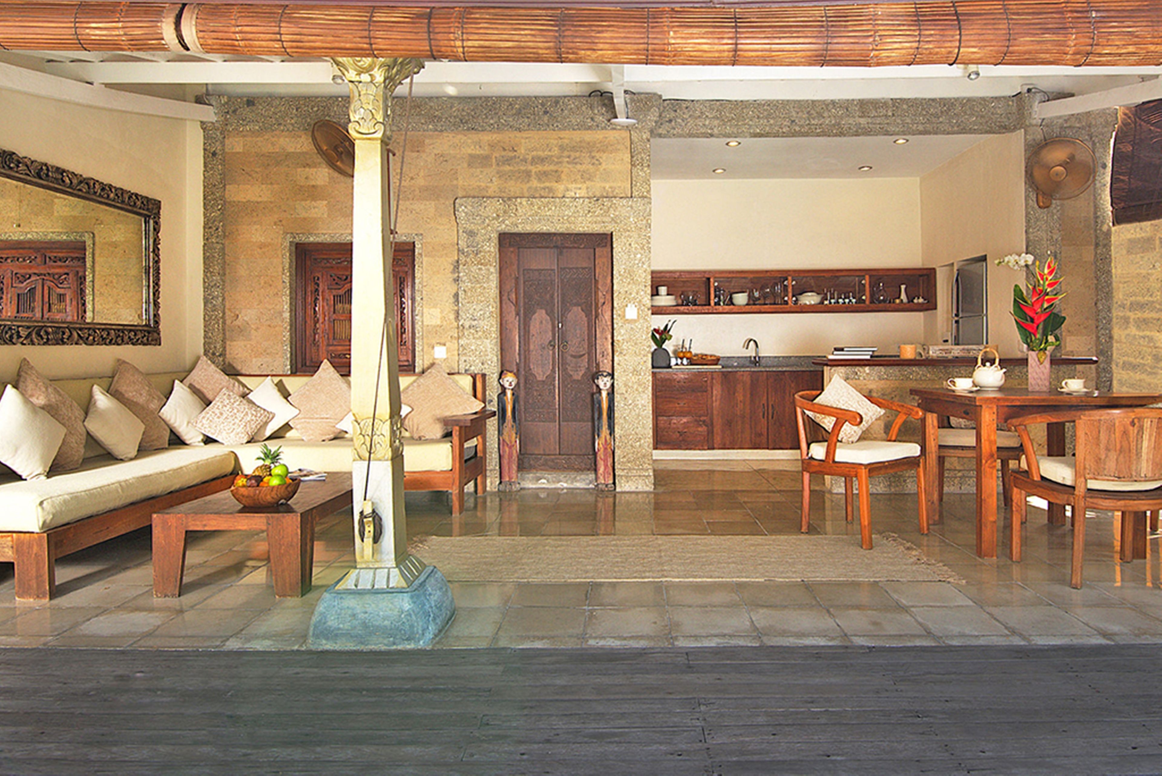 Ampla sala de estar com sofá de madeira, mesa de centro e mesa de jantar com cadeiras. Ao fundo, cozinha com armários de madeira e pia.
