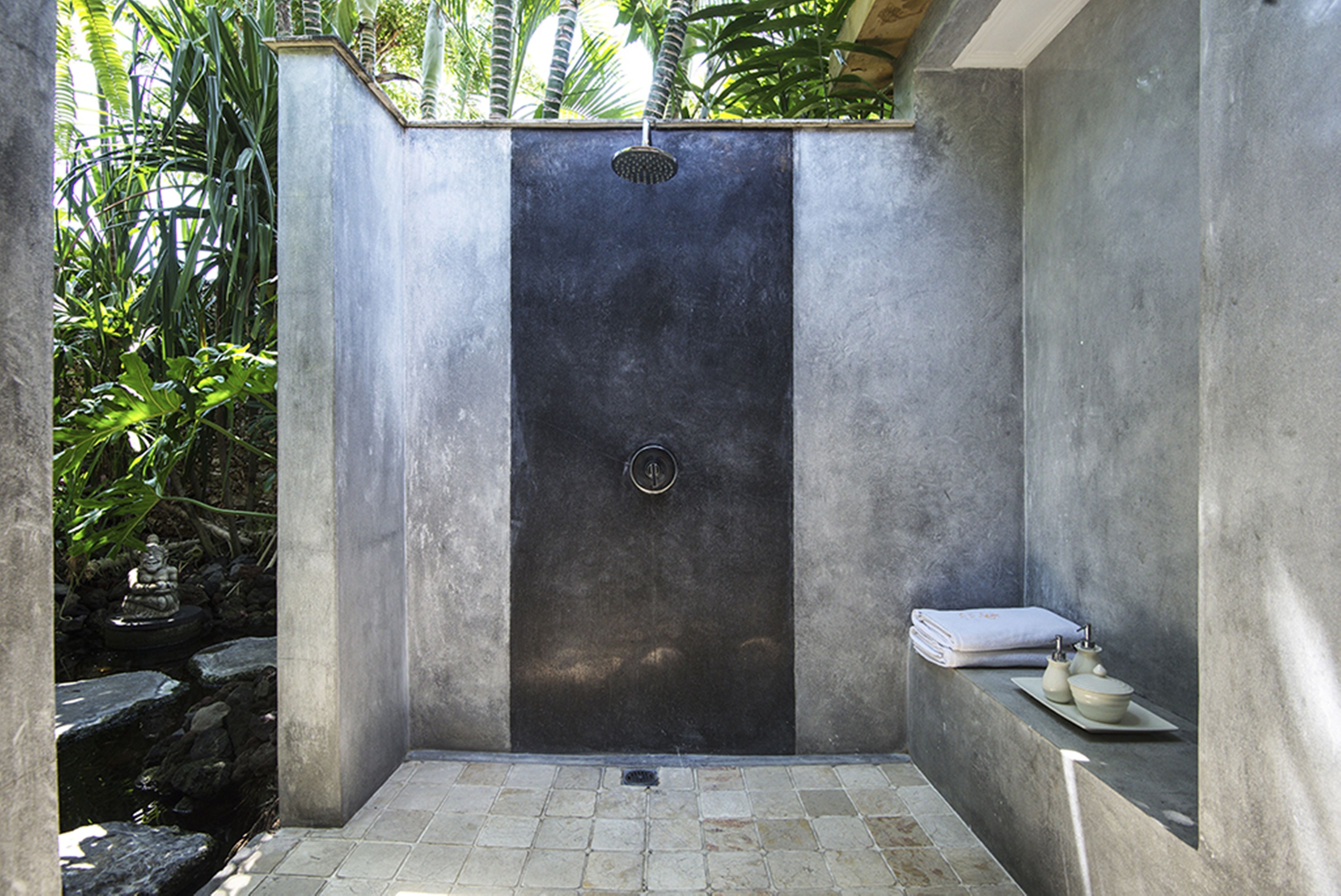 Eine moderne Duschkabine mit dunklem Innenraum, ein kleines Waschbecken mit Handtuch und ein steinbelagter Weg zu einem Wasserspiel.