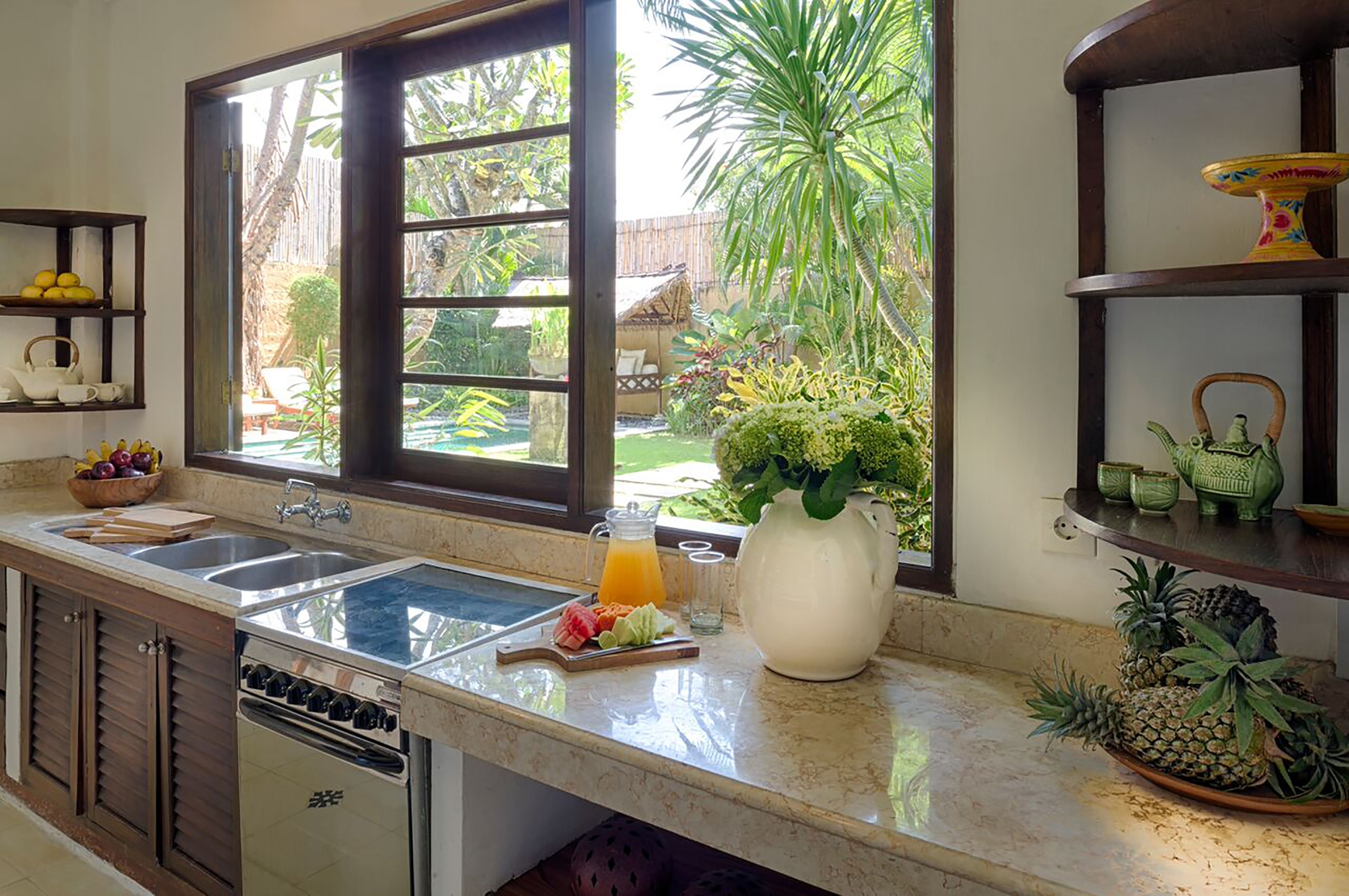 A kitchen with a large window, a sink, a stove, and a countertop with a pitcher of orange juice and a bowl of fruit.