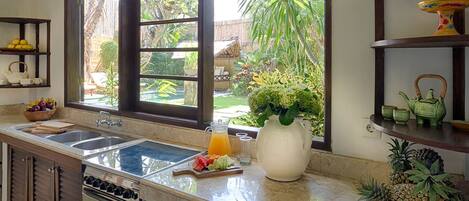 A kitchen with a large window, a sink, a stove, and a countertop with a pitcher of orange juice and a bowl of fruit.