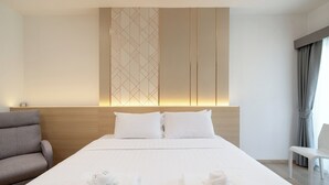 A hotel room with a large bed, two pillows, a headboard with a geometric pattern, a grey armchair, a white chair, and a window with curtains. - Riski Residence (Nonthaburi)
