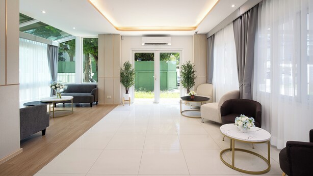 A modern living room with a sofa, armchairs, and a coffee table. There is a large window with curtains, a door leading outside, and a potted plant. - Riski Residence (Nonthaburi)