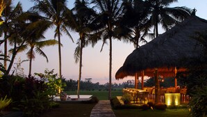 A thatched-roof outdoor dining area with tables and chairs, surrounded by palm trees and a well-maintained lawn. - MATHIS Retreat Ubud (Ubud)