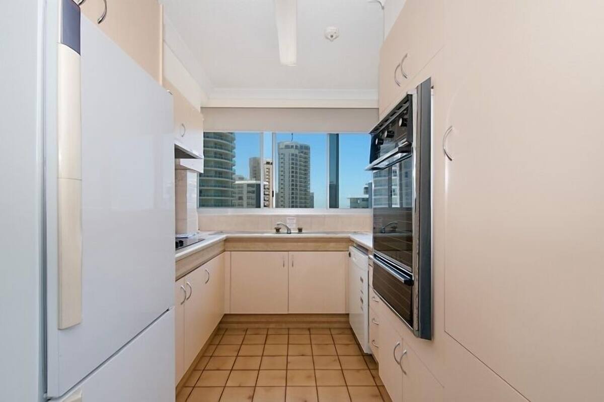 Cocina compacta con armarios blancos, piso de baldosas y vista a edificios altos por una ventana.