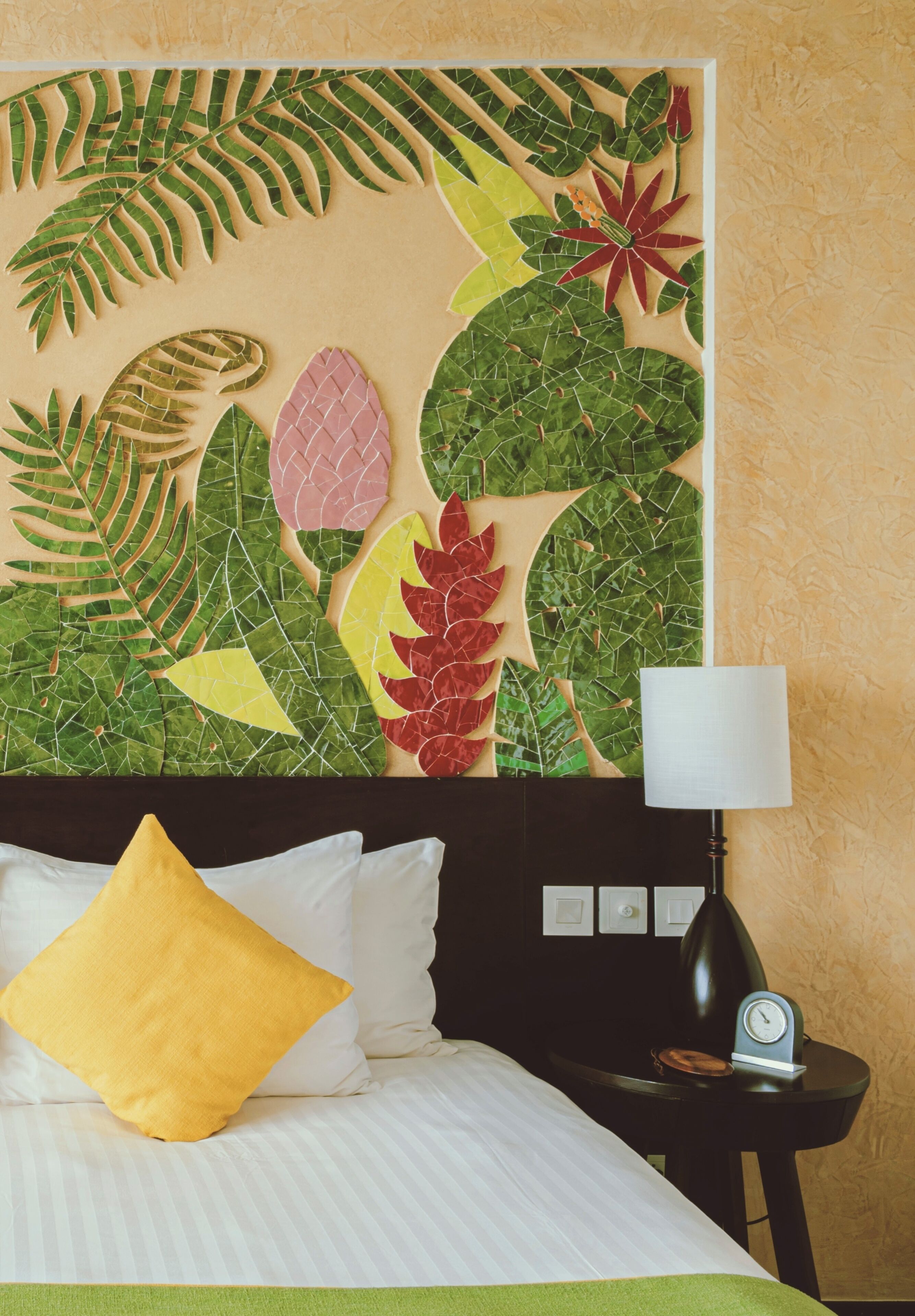 A bed with white bedding and a yellow pillow, a nightstand with a lamp and clock, and a wall with a tropical leaf mosaic.