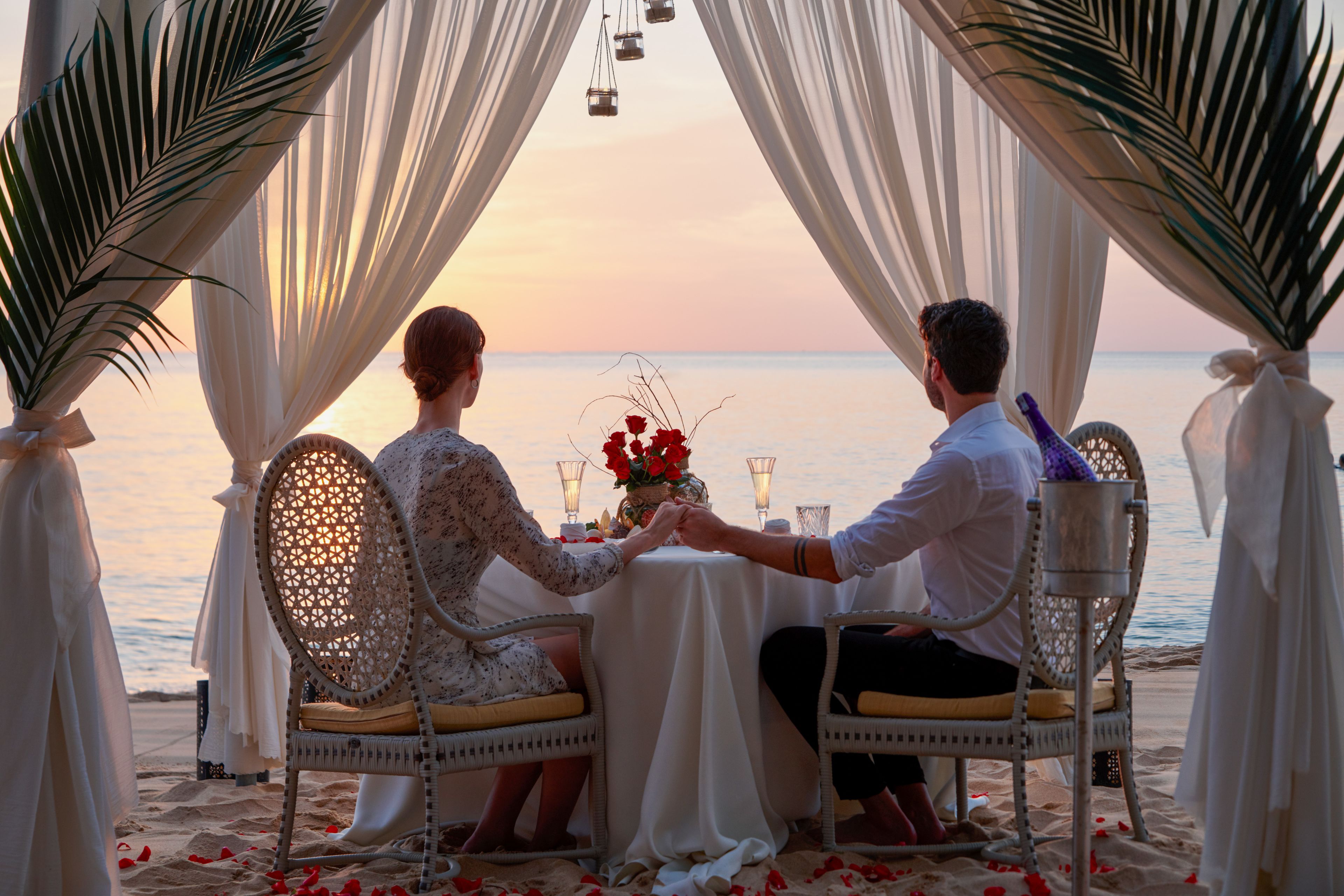 Una pareja sentada a una mesa con vistas al mar, rodeada de telas colgantes y hojas de palmera.