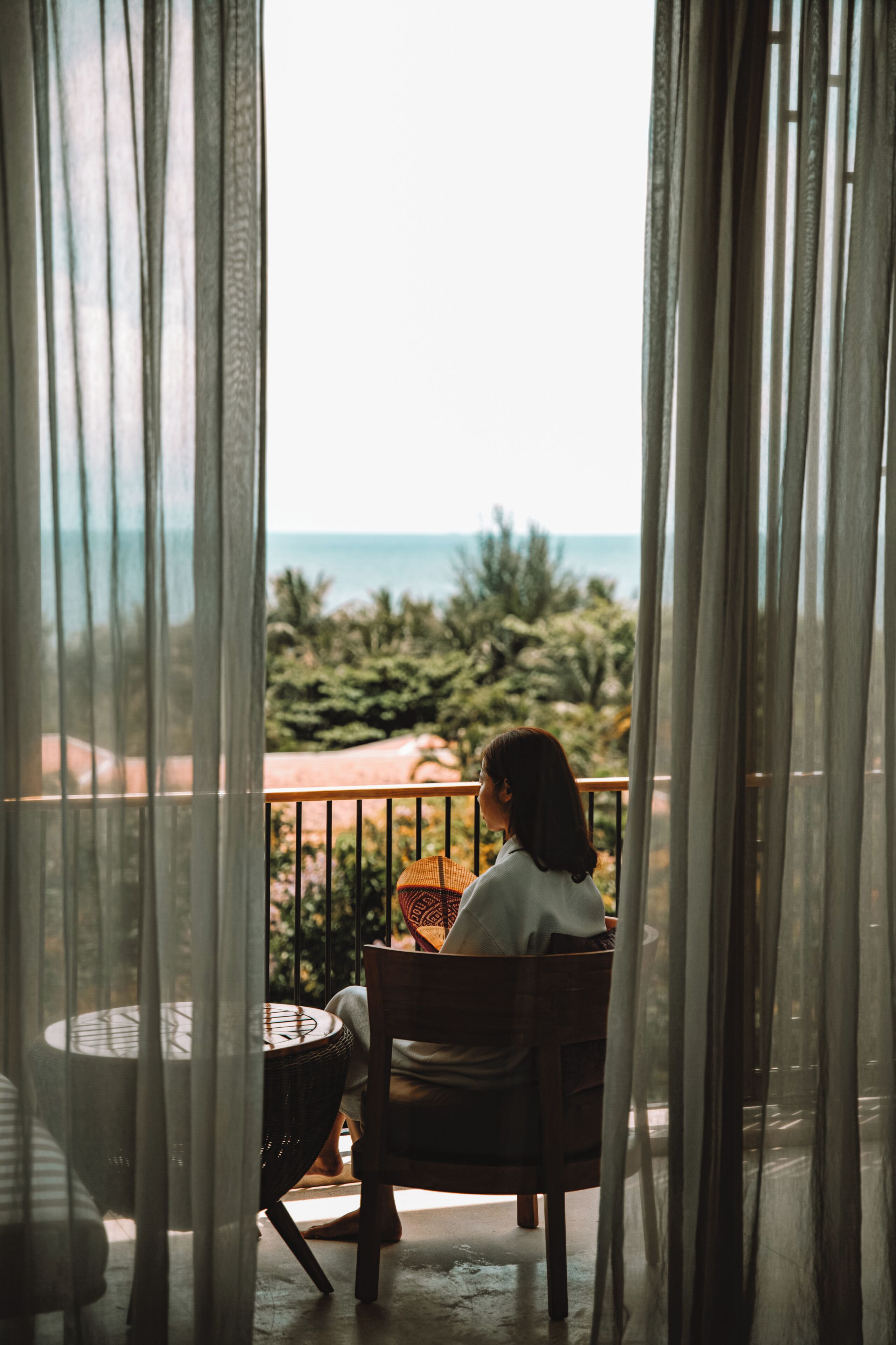 Una persona sentada en una silla de madera en un balcón con vista al mar y follaje.
