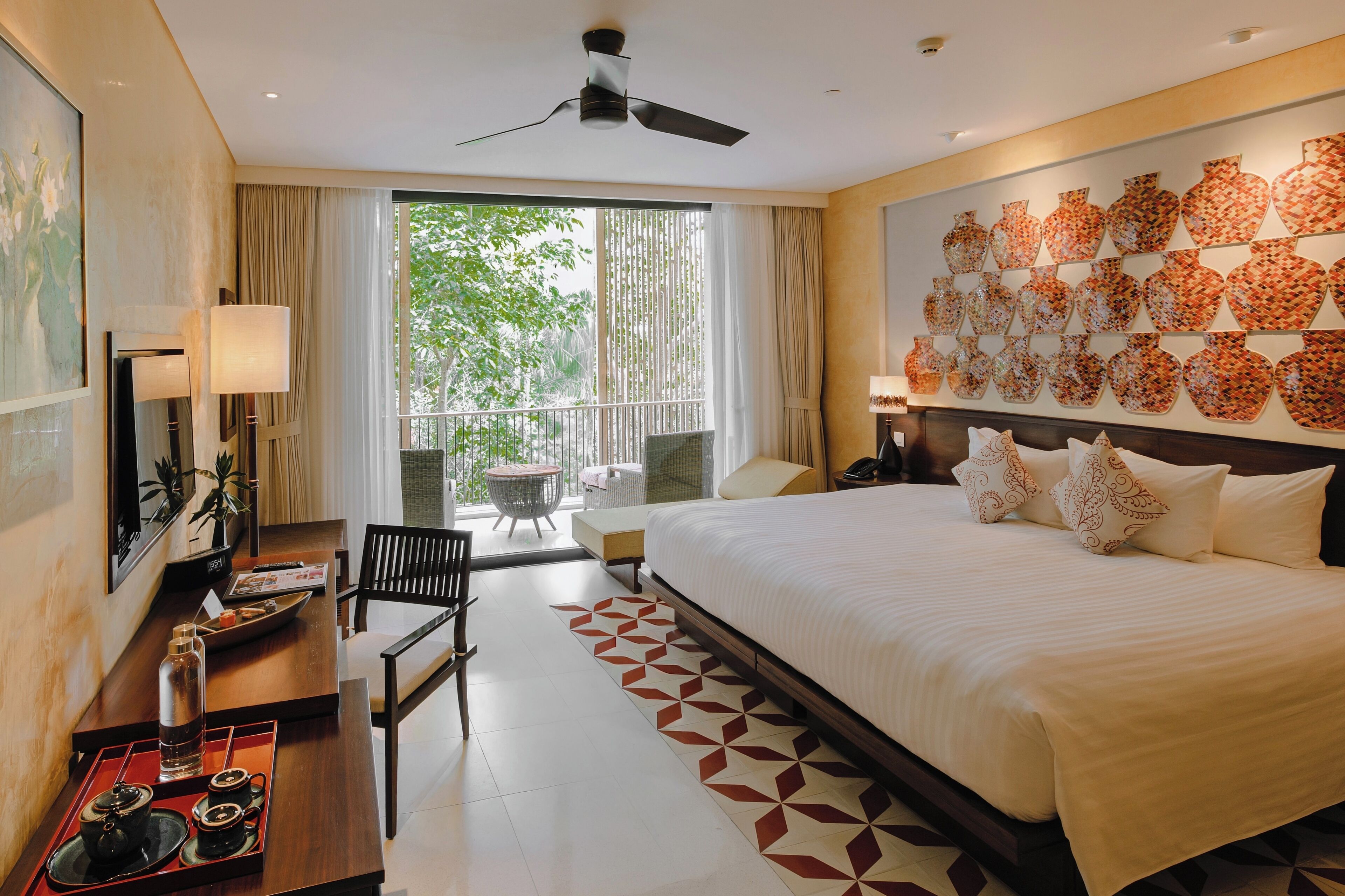 A hotel room with a large bed, a desk with tea set, a balcony with a table and chairs, and a wall with patterned vases.