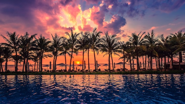 Un atardecer sobre una piscina con palmeras y sombrillas de playa.