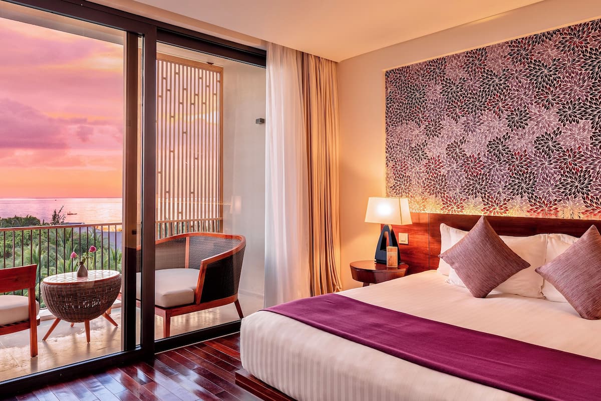 Ein Hotelzimmer mit Bett, Nachttisch, Lampe, Stuhl, kleinem Tisch und Meerblick bei Sonnenuntergang.