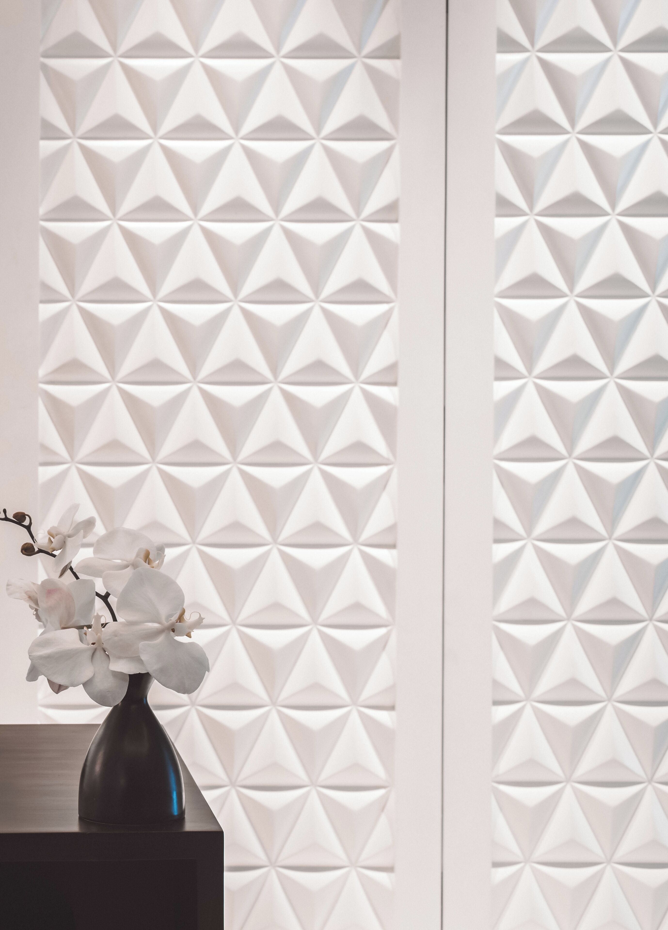 A room with a geometric-patterned partition, a dark-colored vase with white flowers, and a dark wooden surface.