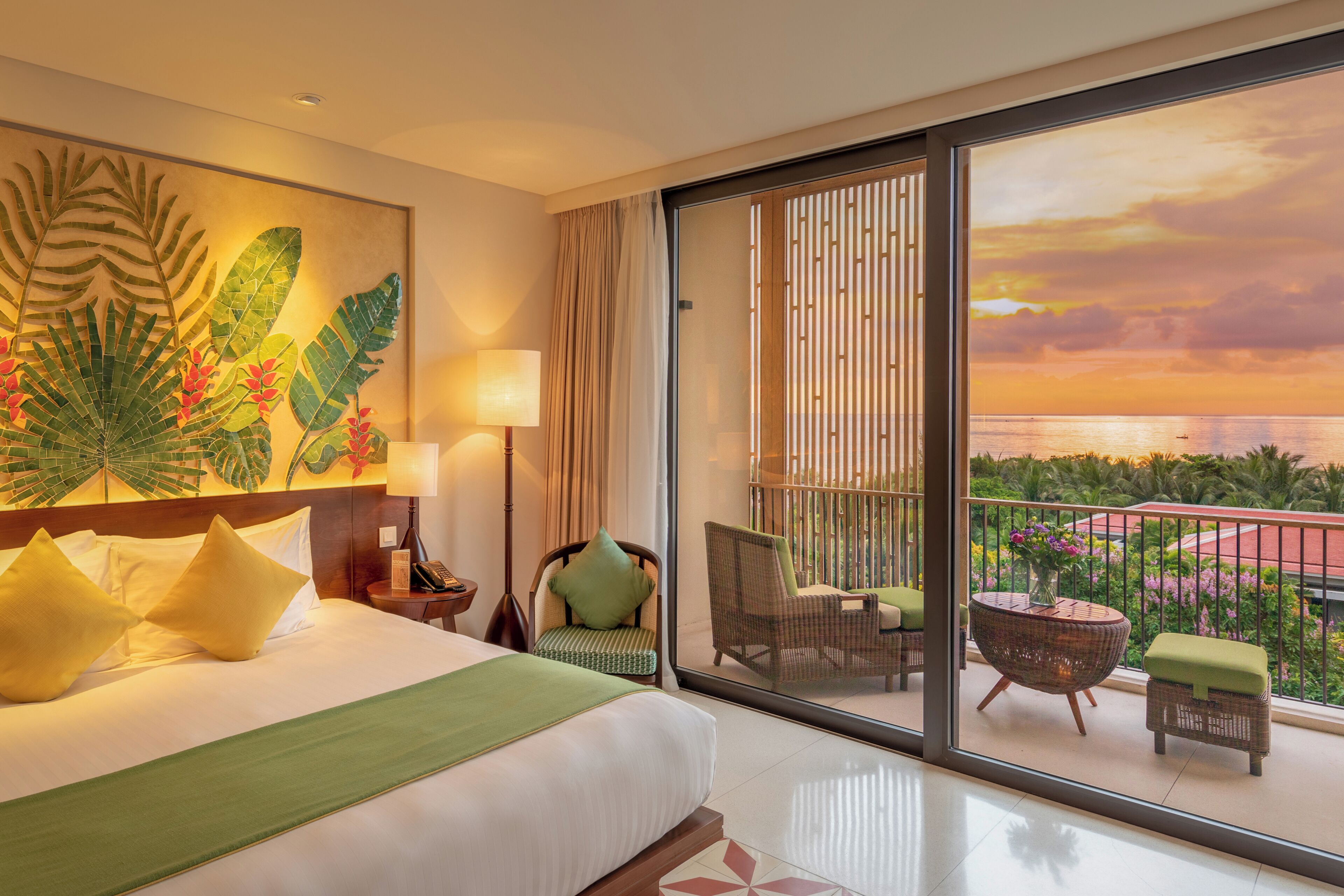 Une chambre d’hôtel avec un lit, une vue sur le coucher de soleil, un balcon avec des chaises et une décoration murale sur le thème des tropiques.