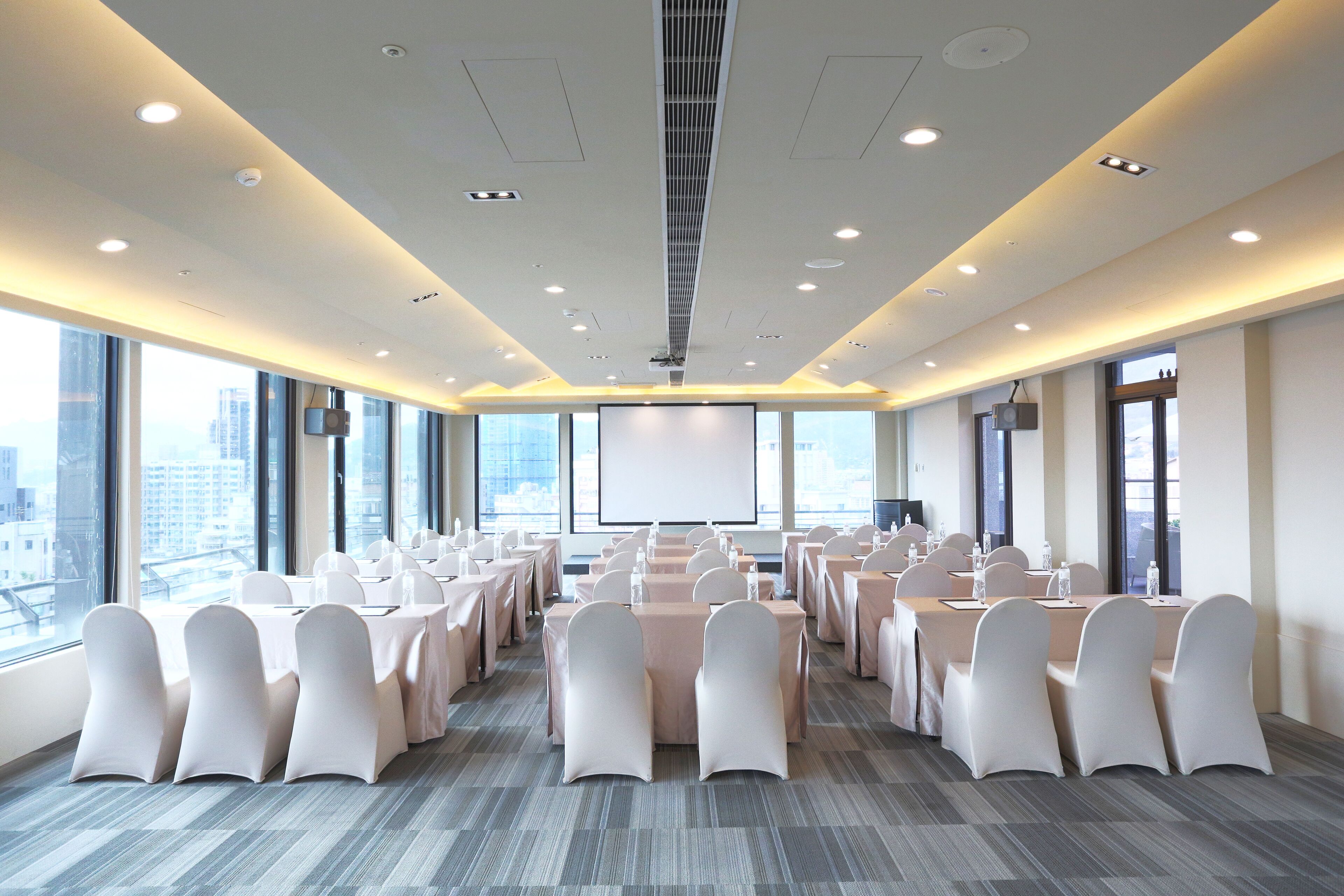 A conference room with rows of tables and chairs, large windows, and a projector screen.