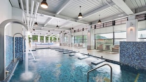 En indendørs swimmingpool med krystalklart vand, flisegulv og udsigt til det fri gennem store vinduer.