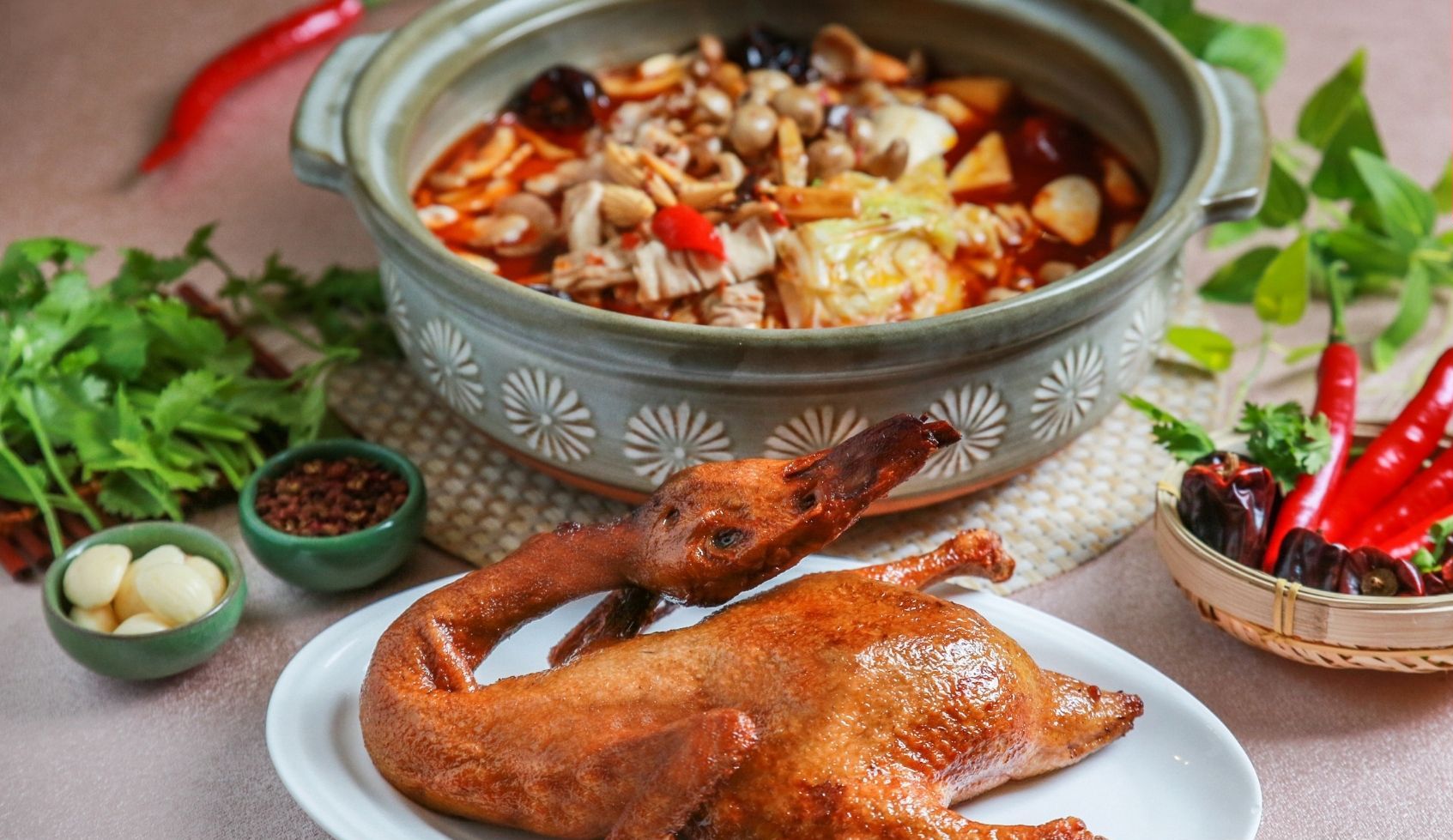 A roasted duck on a plate, a pot of vegetables, and a basket of chili peppers.