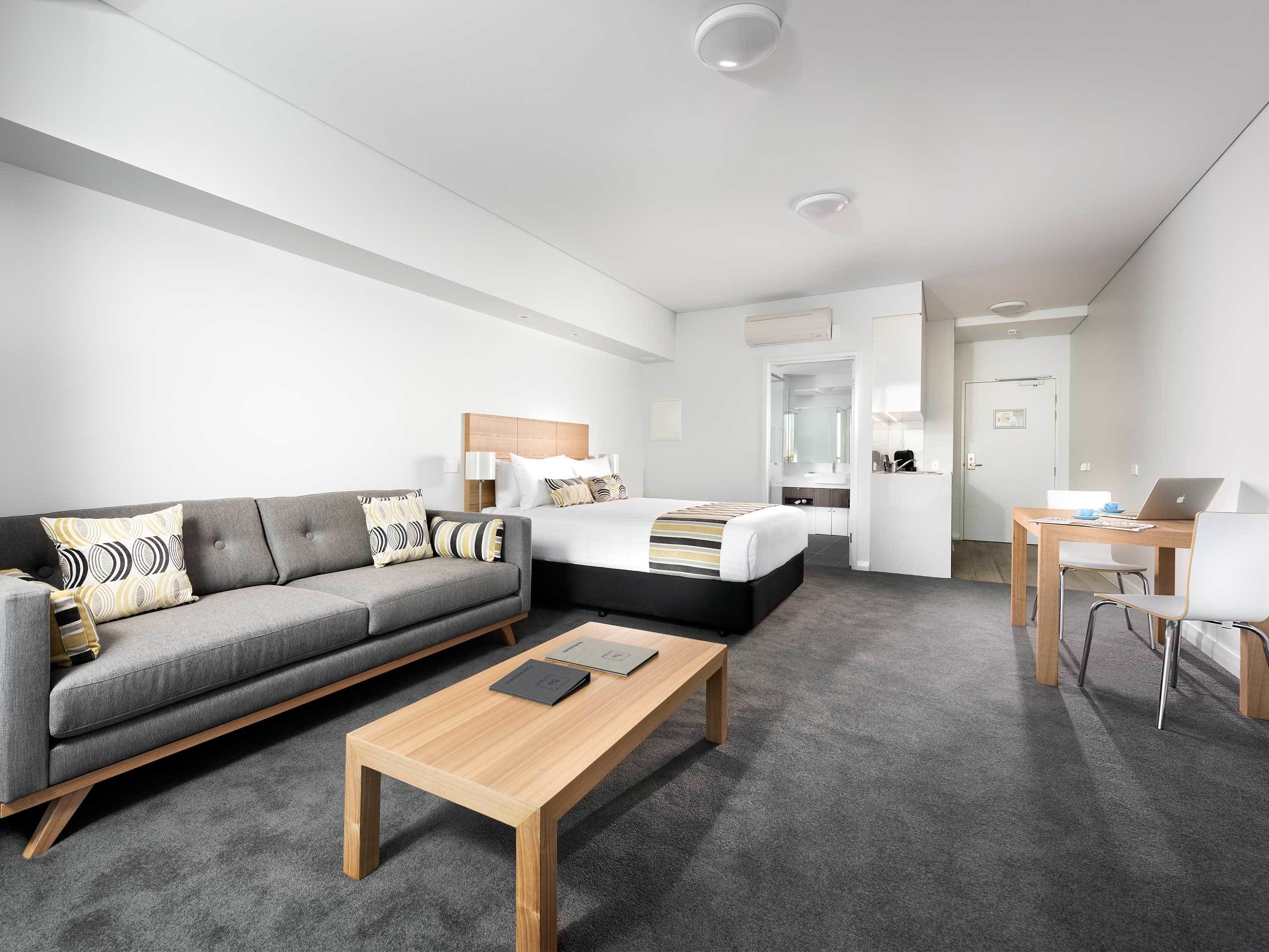 A modern hotel room with a grey sofa, a wooden coffee table, a bed with a striped bedspread, a desk with a laptop, and a bathroom visible through an open door.