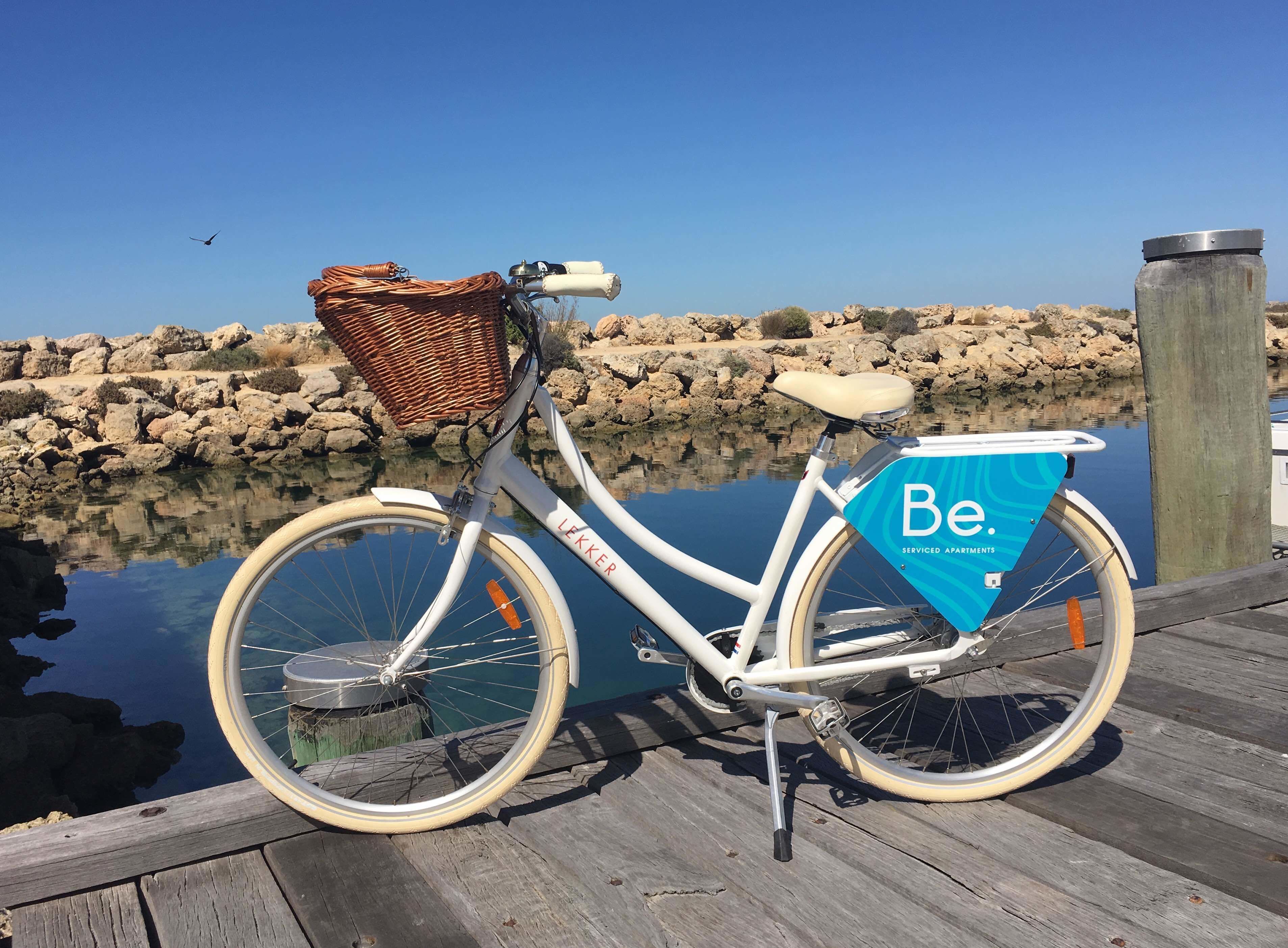 Una bicicleta blanca con canasta de mimbre y una bolsa azul con la etiqueta 'Be. Serviced Apartments' estacionada en un muelle de madera junto a un cuerpo de agua tranquilo.