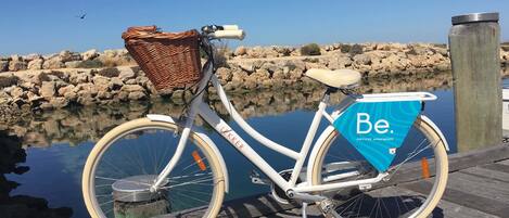 Una bicicleta blanca con canasta de mimbre y una bolsa azul con la etiqueta 'Be. Serviced Apartments' estacionada en un muelle de madera junto a un cuerpo de agua tranquilo.