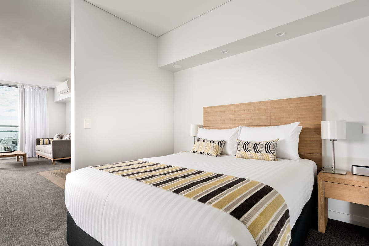 Una habitación de hotel moderna con una cama grande, un cabecero de madera, una mesita de noche con una lámpara y vistas a la ciudad a través de amplias ventanas.