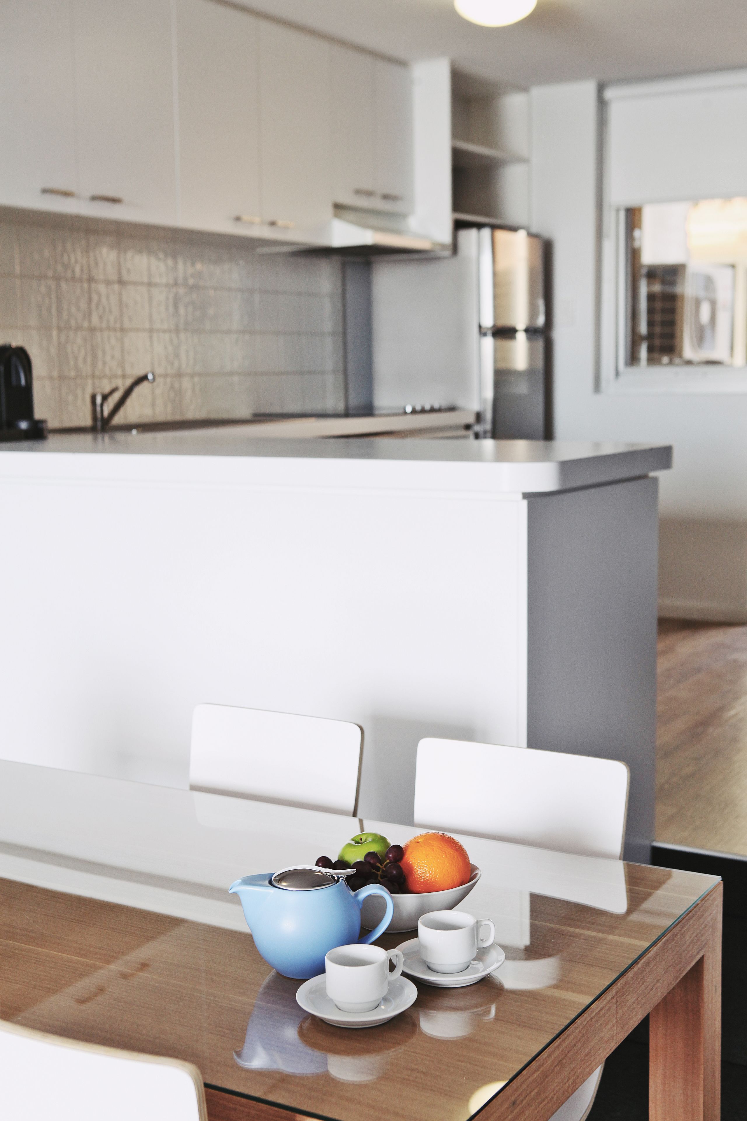 Una cocina moderna con armarios blancos, un refrigerador de acero inoxidable y una mesa de comedor de vidrio con una tetera azul, tazas blancas y un bol de fruta.