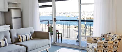 Una sala moderna con un sofá, un sillón estampado, una mesa de centro con un frutero y vista al mar a través de amplios ventanales correderas.