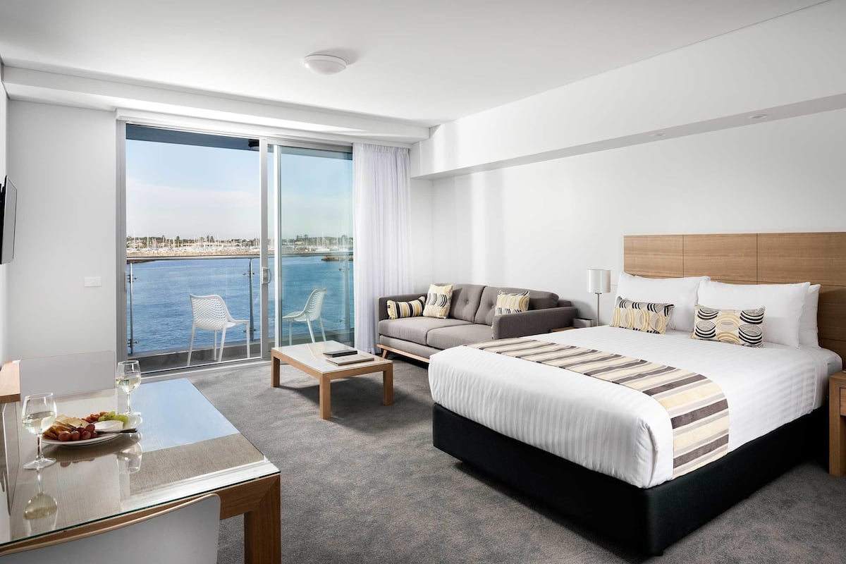 Habitación de hotel con cama, sofá, mesa de comedor con superficie de vidrio y vista al mar.