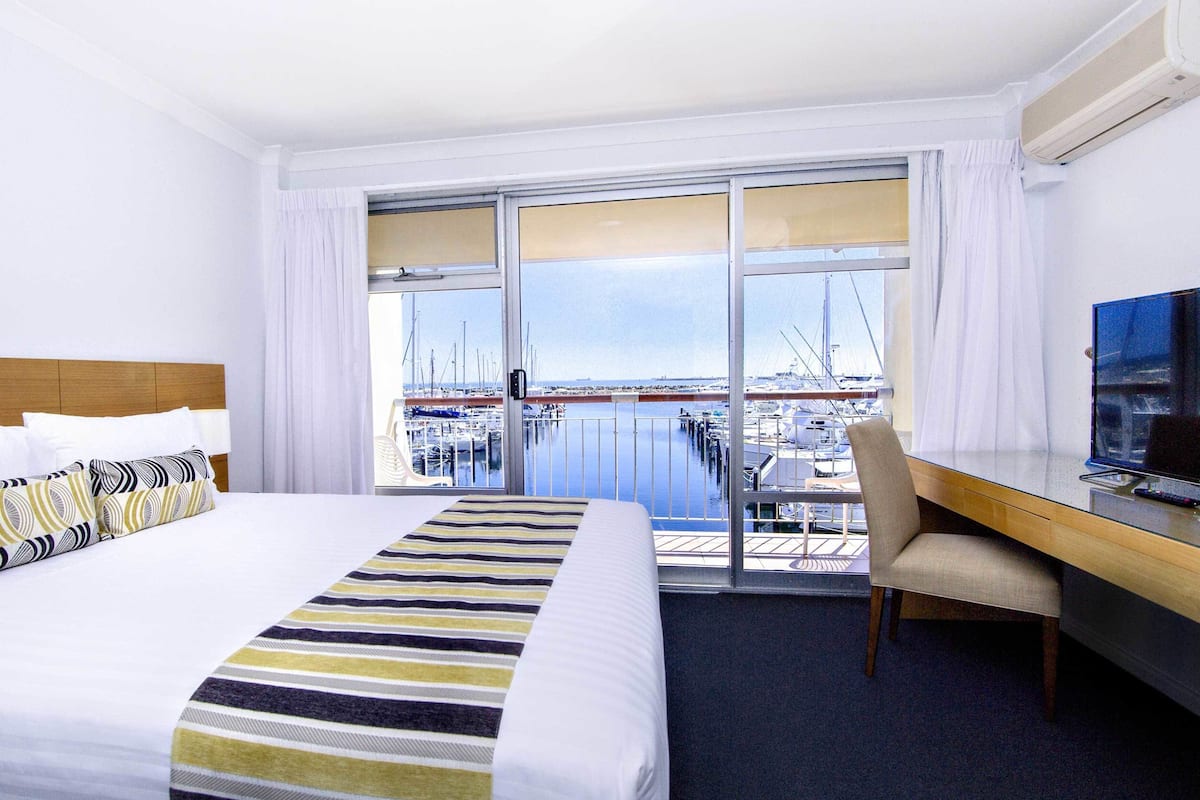 Habitación de hotel con cama, escritorio, silla y televisor. La habitación tiene vista a un puerto deportivo con barcos.