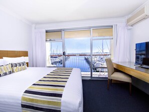 A hotel room with a bed, a desk, a chair, and a TV. The room has a view of a marina with boats.