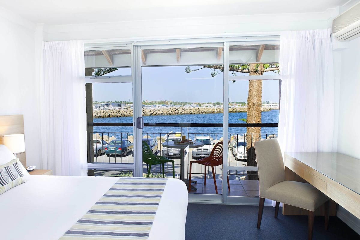Habitación de hotel con cama, escritorio, silla y balcón con vista al mar y coches estacionados.