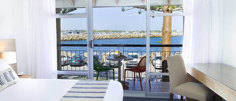 Een hotelkamer met een bed, een bureau, een stoel en een balkon met uitzicht op zee en geparkeerde auto's.