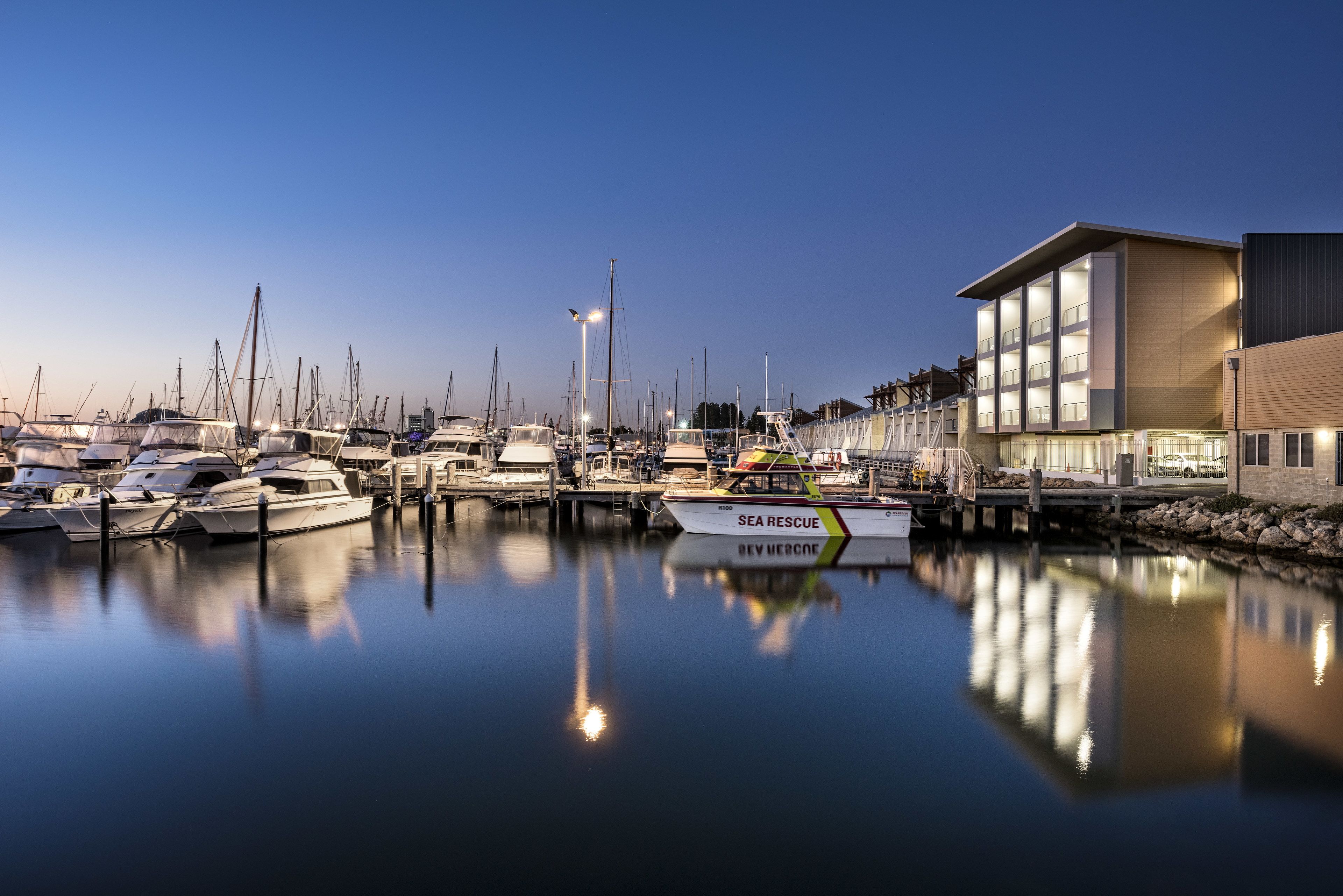 A marina with boats docked, a modern building with large windows, and a calm water surface reflecting the boats.