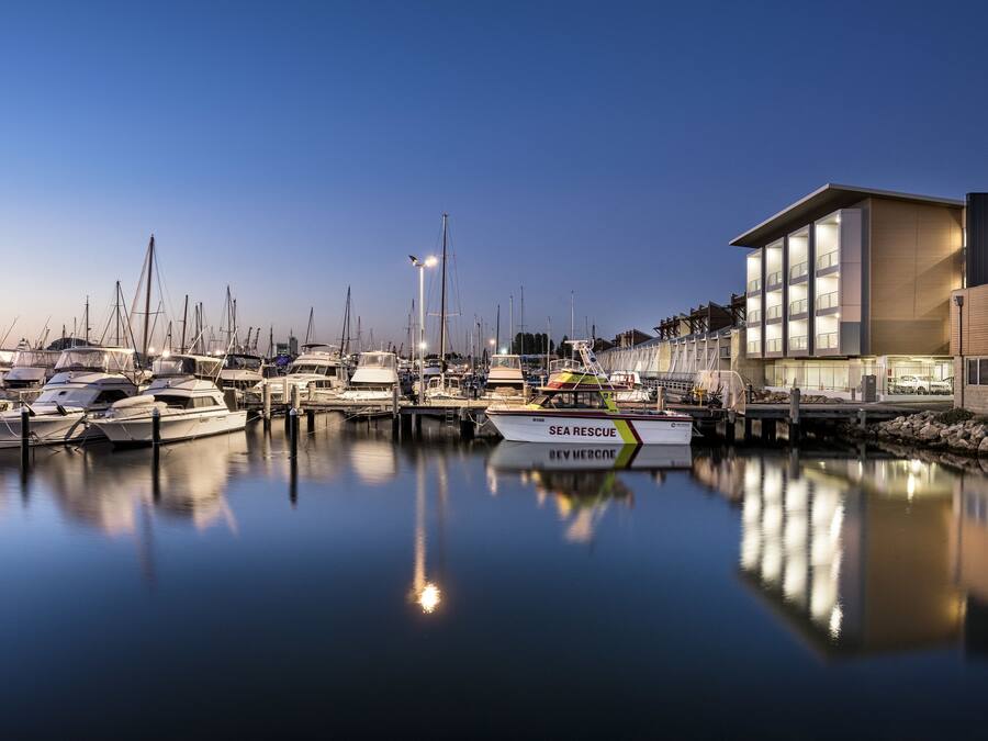 A marina with boats docked, a modern building with large windows, and a calm water surface reflecting the boats.