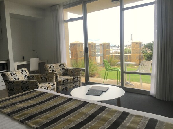 A modern hotel room with a large window, a sofa, a dining table, and a patterned rug.