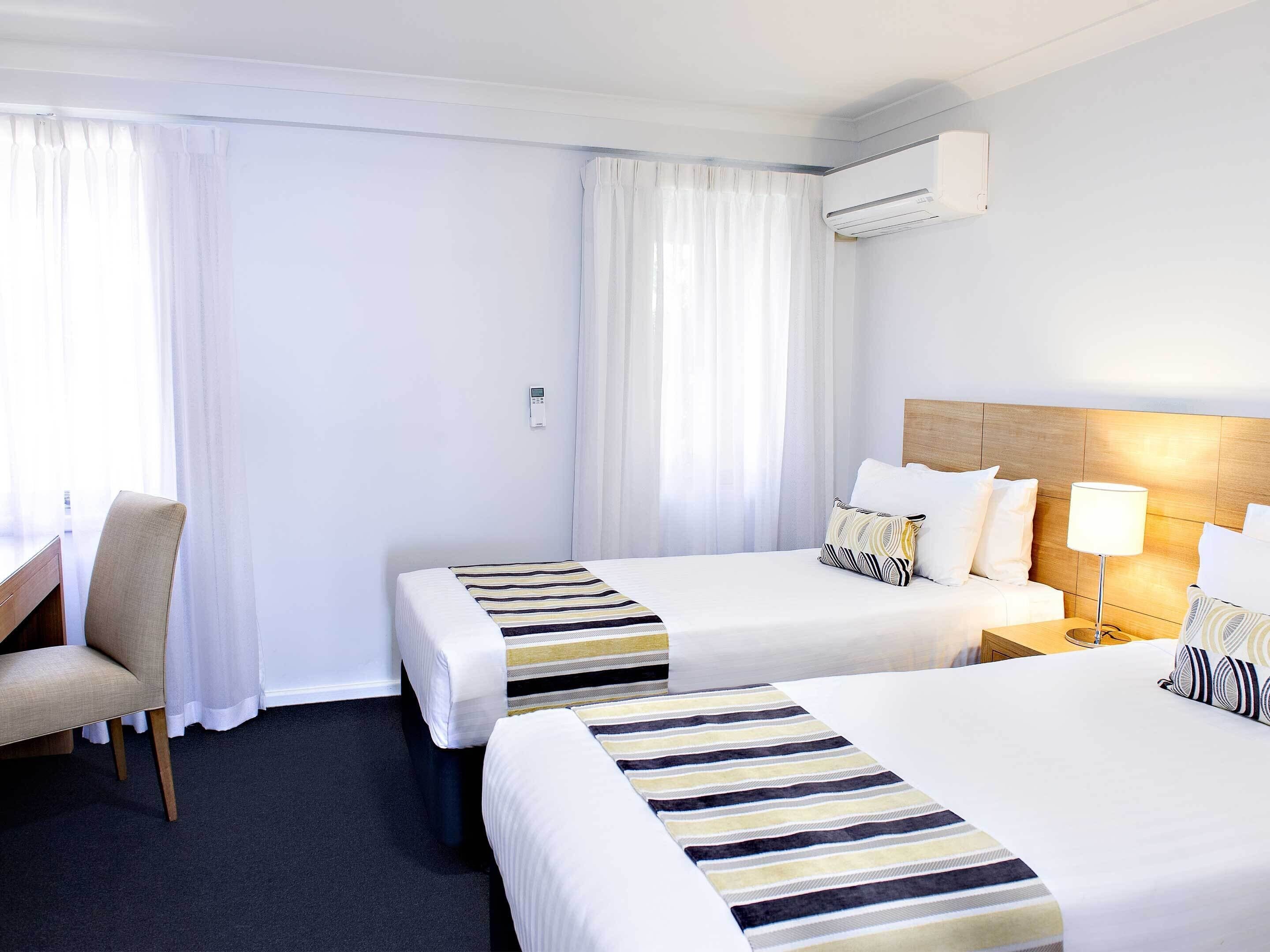 A hotel room with two beds, a desk, a chair, a lamp, and an air conditioning unit.