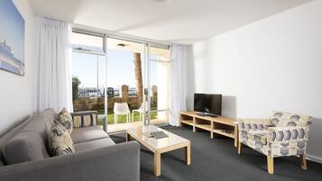 A modern living room with a sofa, coffee table, and patterned armchair. A television is on a wooden stand, and there is a large window with curtains.