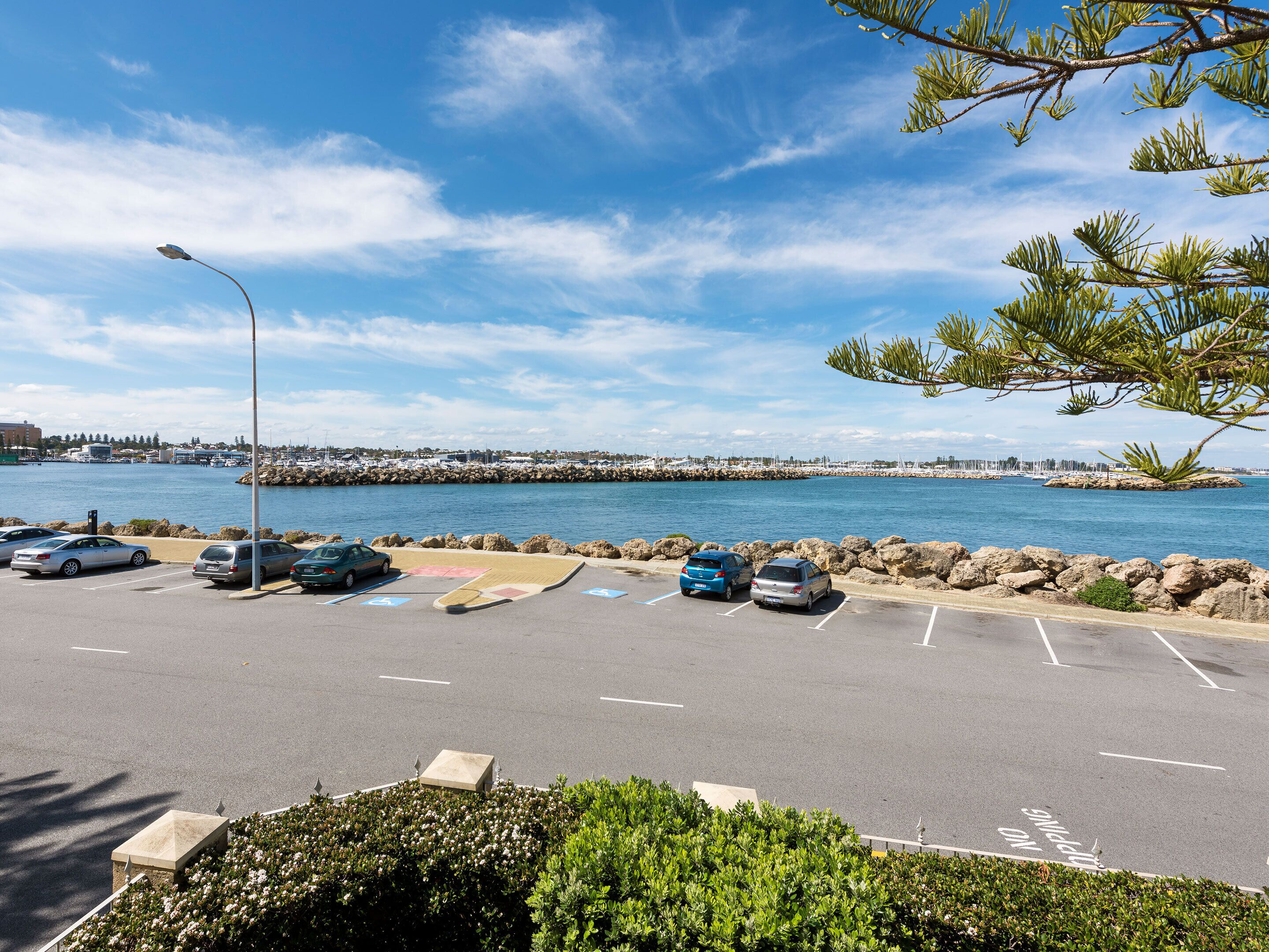 Un estacionamiento con coches, cielo despejado y un puerto deportivo al fondo.