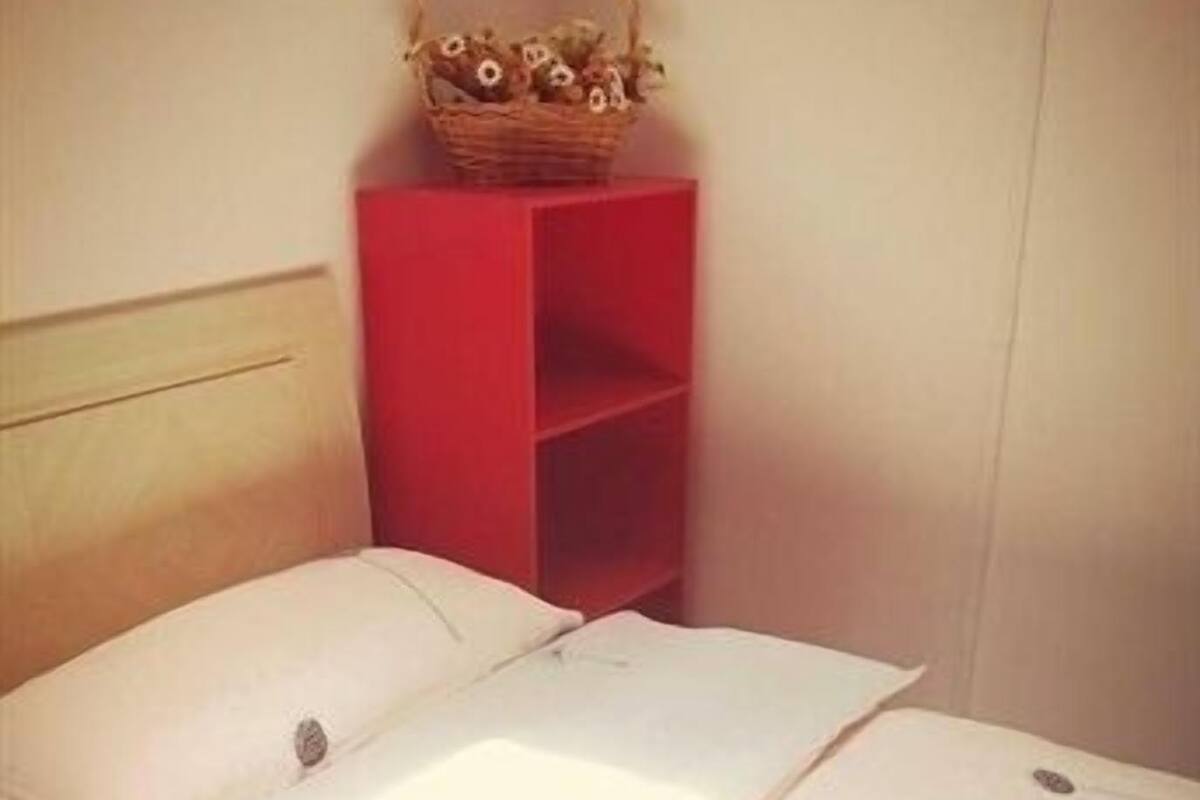 A bed with white linens and pillows, a red nightstand with a basket, and a wooden headboard.
