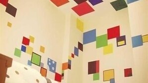 Una habitación con una cama, una pared con formas geométricas coloridas y una ventana con vista al cielo.