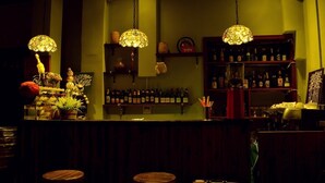 Una zona de bar con mostrador de madera, lámparas colgantes, estantes con botellas y taburetes.