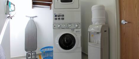 Uma lavanderia com máquina de lavar e secadora empilhadas, um cesto de roupa azul e um bebedouro.