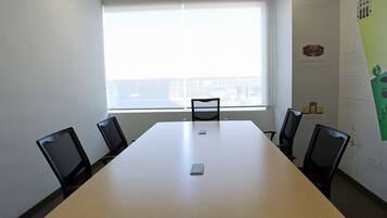 Un salón de conferencias con una mesa grande, cuatro sillas, un ventanal con persianas y una pared con carteles informativos.