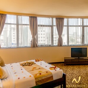 A hotel room with a bed adorned with rose petals, a television on a stand, and large windows with curtains. - Nazra Hotel (Addis Ababa)