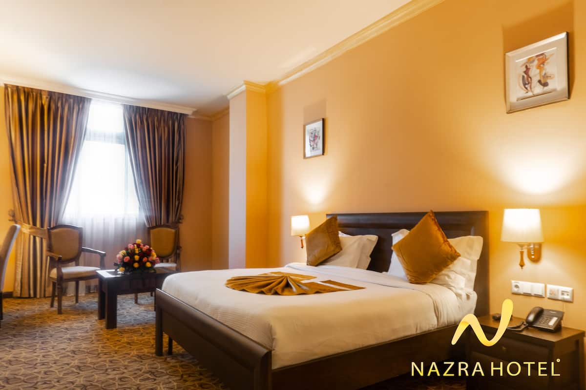 A hotel room with a bed, two bedside lamps, a nightstand with a phone, a desk with a lamp, a chair, and a window with curtains.