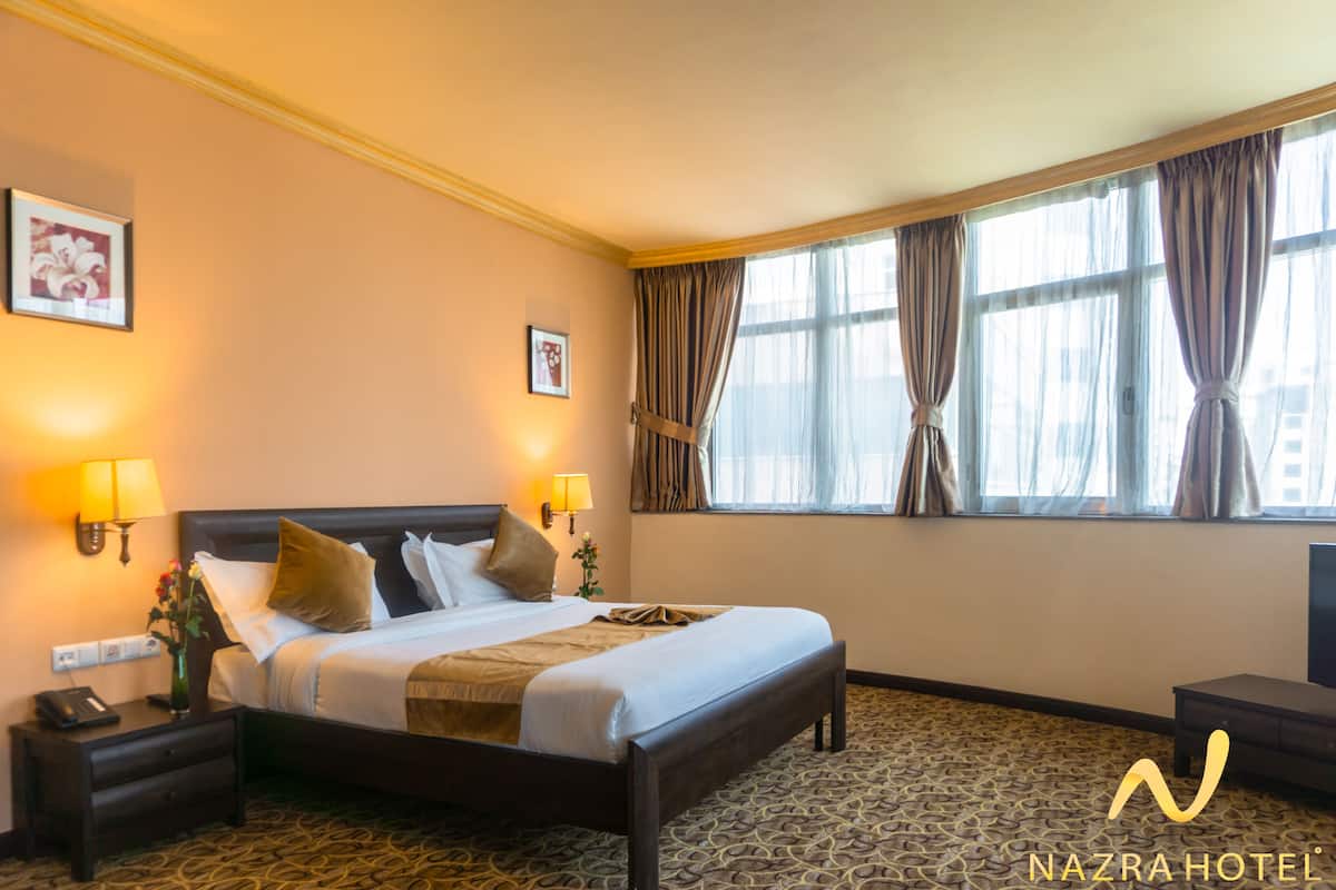 A hotel room with a large bed, two bedside lamps, a nightstand, a window with curtains, and a carpeted floor.